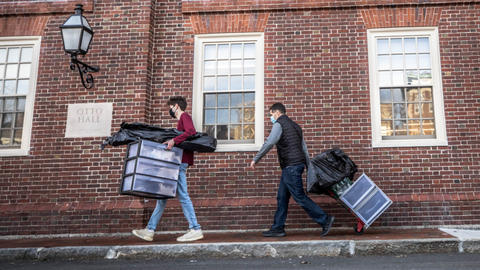 Two people walking alongside a building, one carrying boxes and bags and the other pulling a cart with boxes and bags