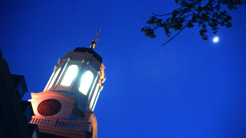 Skyward view showing an illuminated belfry against a darkening sky with a gibbous moon