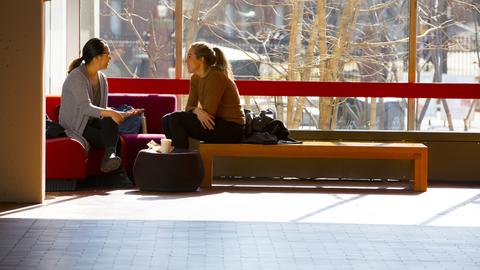 Two people talking in a room corner in front of a large picture window.