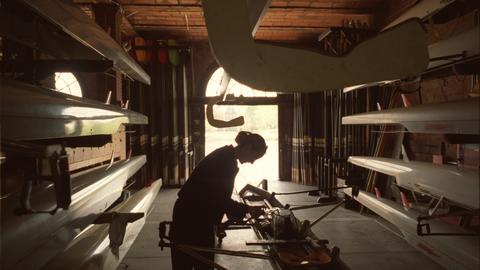 Person working on a crew boat inside a boat house, framed by rows of crew boats.