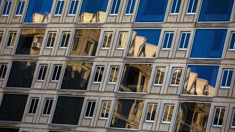 Building exterior showing a grid of paired windows and the reflection of an adjacent building and blue sky.