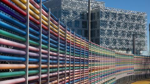 Curved fence with rails of alternating pride colors in front of a modern geometric building.