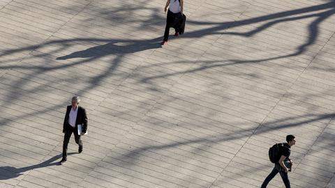 View from above of plaza with tree branch shadows and three people walking alone in different directions.