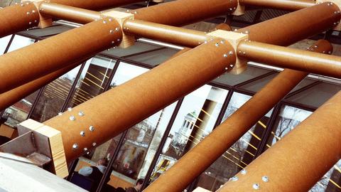 Closeup of a structure of tubes connected to wooden blocks, overlaying a window and buildings beyond.