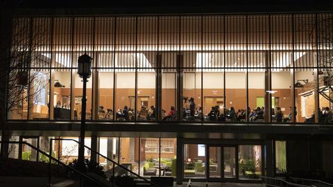 Night view of a building with people visible inside through large glass windows, attending a gathering or lecture.
