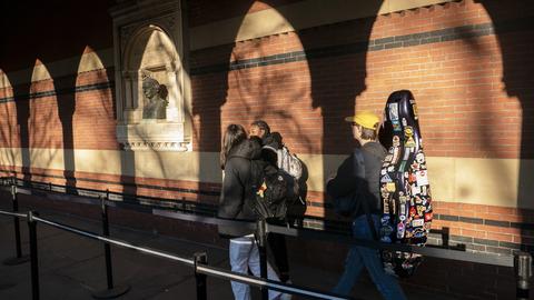 People walking past a brick wall carrying backpacks and a sticker-covered cello case. Shadows of arches are cast on the wall.