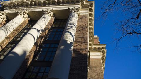 A classical building with ornate columns and a brick facade, with tree shadows cast against it under a clear blue sky.