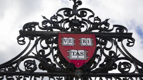 Ornate black wrought iron gate featuring a Veritas shield and a cloudy sky in the background.